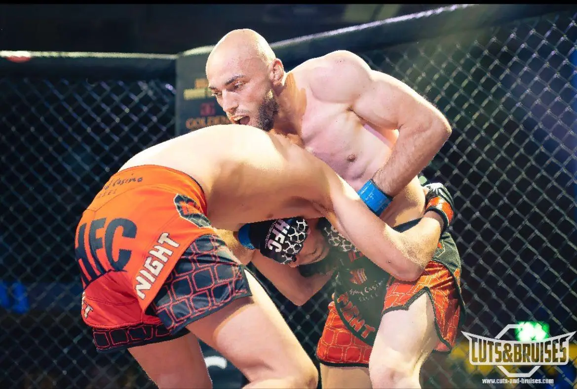 Combat de MMA en cage : un combattant au crâne rasé contrôle son adversaire en clinch.