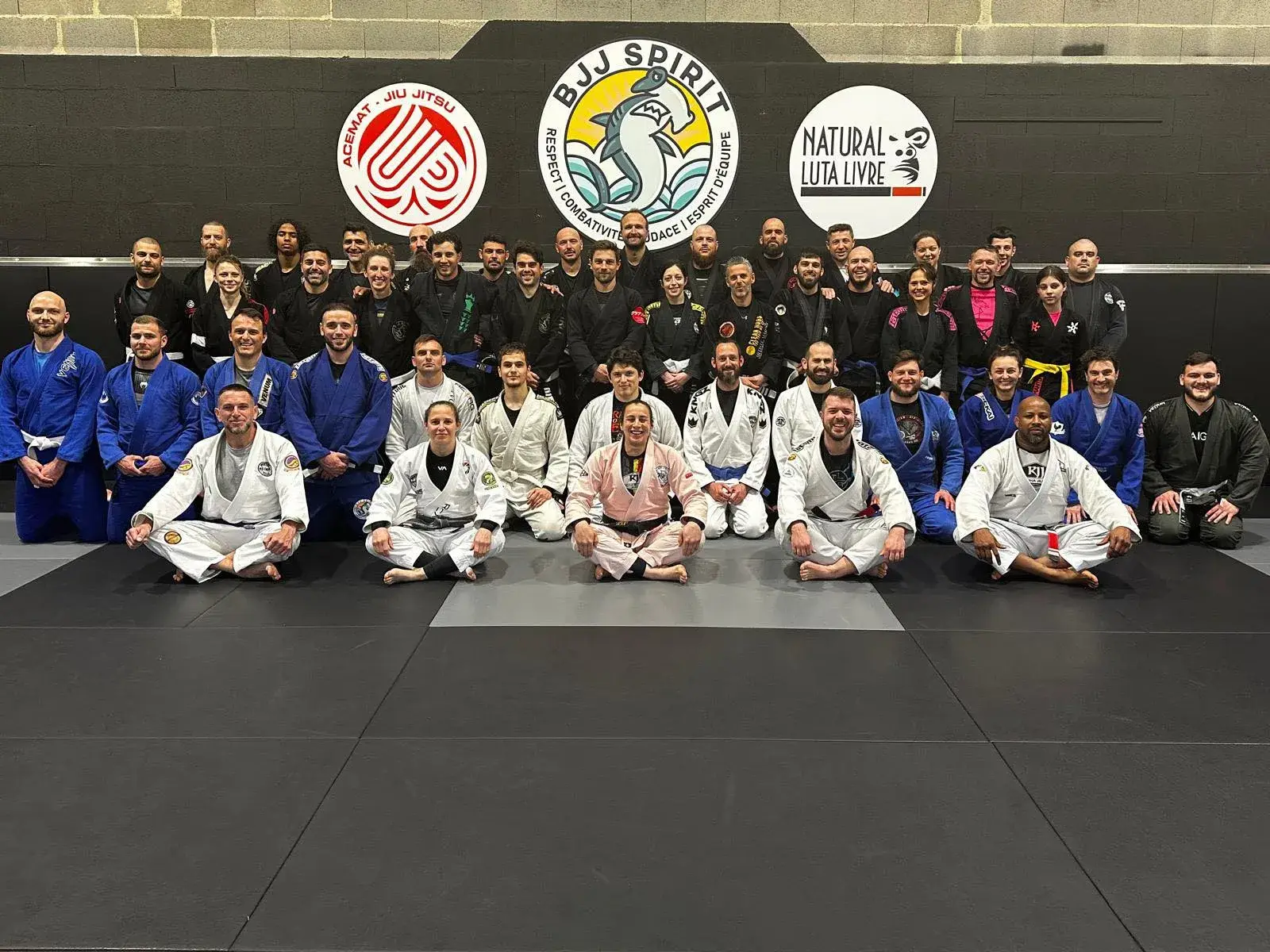 Photo de groupe sur le tapis de BJJ Spirit, devant les logos Acemat, BJJ Spirit et Natural Luta Livre peints au mur.